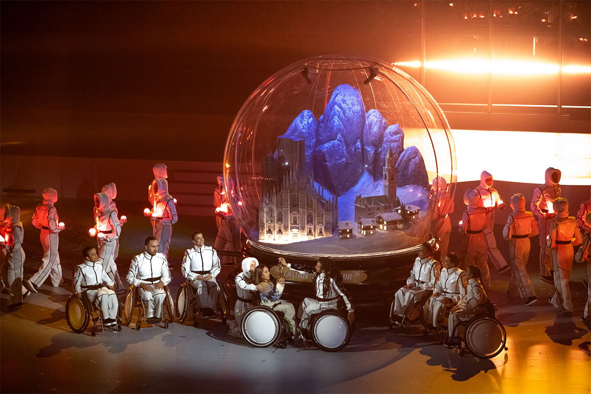 Lavalla tanssijoiden ympäröimänä suuri matkamuisto-lumisadepallo, jonka sisällä Milanon ja Cortinan kaupunkien maamerkkirakennuksia.